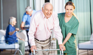 An elderly man using a walker is assisted by a smiling healthcare worker in green scrubs, with two other elderly individuals interacting in the background.