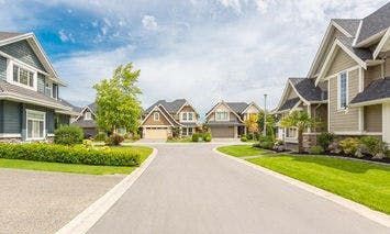 Suburban street lined with modern, detached houses featuring gabled roofs and manicured lawns on a sunny day, with a clear blue sky and scattered clouds in the background.