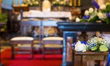 Flowers rest on a pew beside an altar adorned with candles and flowers, with wooden benches in a church featuring a red carpet aisle.