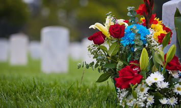 A bouquet of colorful flowers rests against a gravestone in a cemetery, surrounded by a field of green grass and rows of other gravestones extending into the blurred background.