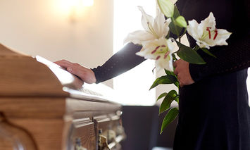 Person in black clothing holding a bouquet of white lilies, resting one hand on a wooden casket, inside a softly lit room.