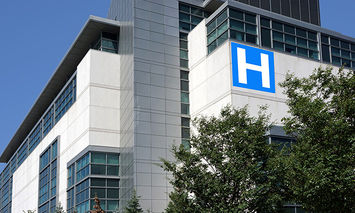 A tall, modern hospital building with reflective windows displays a large blue 