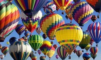 Numerous brightly colored hot air balloons float upwards against a clear blue sky, creating a vibrant, uplifting scene with various patterns and designs on each balloon.