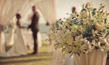 A bouquet of white flowers sits in the foreground while a couple holds hands under a draped canopy in the background, set outdoors in a grassy area.